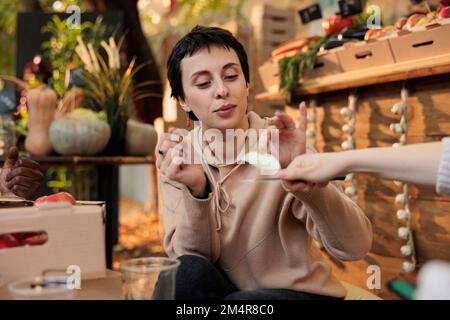 Une cliente de sexe féminin goûtant une tranche de pomme verte biologique fraîche avant de faire ses achats sur le marché agricole extérieur. Le client sourit et goûte des produits biologiques cultivés localement avant d'acheter sur le stand. Banque D'Images