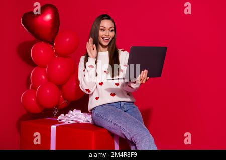 Photo de confiant drôle dame porter blanc chandail assis grand paquet communiquant gadget waving main salutations isolé rouge couleur fond Banque D'Images