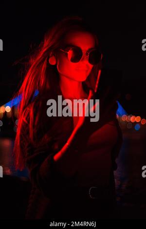Jeune femme d'affaires à la mode portant des lunettes de soleil utilisant un téléphone mobile à l'extérieur avec lumière rouge néon illuminée debout la nuit Banque D'Images