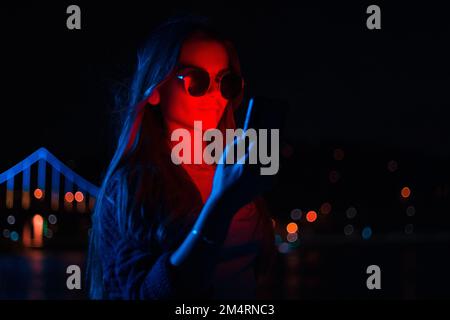 Jeune femme d'affaires à la mode portant des lunettes de soleil utilisant un téléphone mobile à l'extérieur avec lumière rouge néon illuminée debout la nuit Banque D'Images