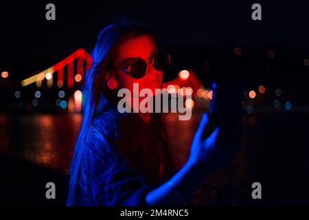 Jeune femme d'affaires à la mode portant des lunettes de soleil utilisant un téléphone mobile à l'extérieur avec lumière rouge néon illuminée debout la nuit Banque D'Images