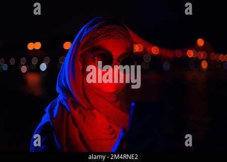 Portrait d'une jeune femme d'affaires musulmane portant des lunettes de soleil en hijab à l'aide d'un smartphone moderne avec lumière rouge néon illuminée son visage la nuit Banque D'Images