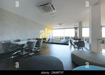 Un salon avec des tables à manger blanches avec des chaises pivotantes grises, de grandes fenêtres et un escalier avec une rambarde en verre Banque D'Images