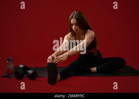 Belle souplesse brunette femelle pull chaussette sur elle-même faisant l'exercice d'étirement après l'entraînement dur. Jolie femme en noir montrant des poses de yoga sur le tapis dans le studio rouge. Concept de mode de vie sain. Banque D'Images