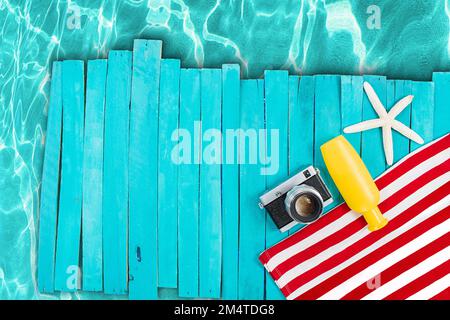 Serviette rayée rouge et blanche avec appareil photo, bouteille jaune, étoile de mer sur des planches en bois bleu, jetée. Piscine, plage, mer. Vertical, horizontal. Vacances, voyages Banque D'Images