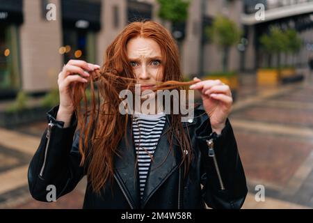 Portrait d'une jeune femme déjumelée touchant des cheveux mouillés après la pluie d'automne, debout sur une belle rue de la ville, regardant l'appareil photo. Banque D'Images