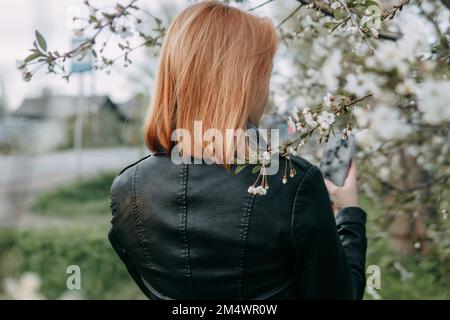 Une adolescente aux cheveux rouges écoute de la musique dans un casque, configuré par téléphone. Moment de la floraison printanière des vergers de cerisiers Banque D'Images