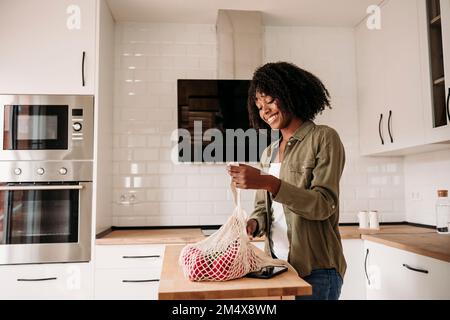 Femme souriante retirant les légumes du sac en filet dans la cuisine à la maison Banque D'Images