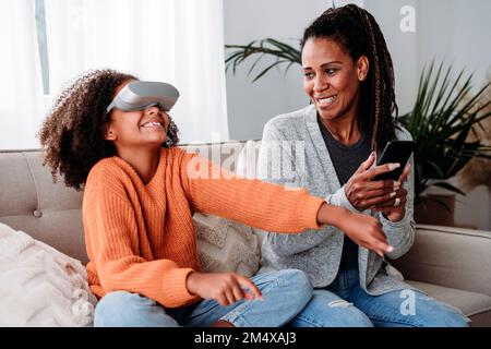Mère souriante avec fille portant un simulateur de réalité virtuelle sur un canapé à la maison Banque D'Images