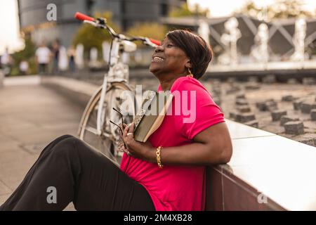 Femme aînée souriante avec livre assis près du mur Banque D'Images