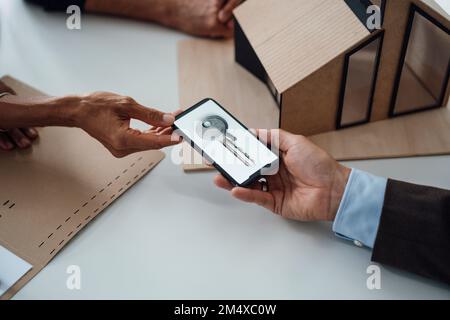 Mains coupées de l'agent immobilier donnant au client un téléphone portable avec la clé de la maison à l'écran Banque D'Images