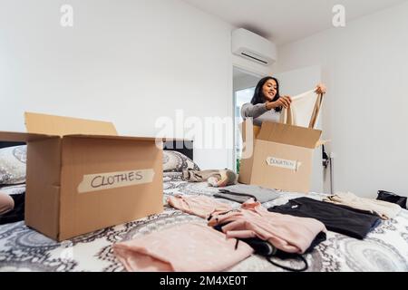 Jeune femme souriante emballant une boîte de vêtements à donner sur le lit Banque D'Images