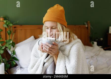 Femme souriante portant un chapeau en tricot qui sent le thé dans la chambre Banque D'Images