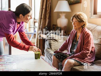 Joyeux soignant qui mélange le café et parle à une femme âgée assise sur un canapé à la maison Banque D'Images