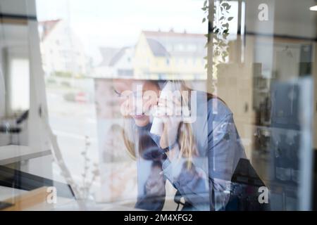 Femme souriante parlant au téléphone vu par une fenêtre en verre dans un salon de coiffure Banque D'Images