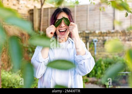 Jeune femme gaie tenant les feuilles par-dessus les yeux dans le jardin Banque D'Images