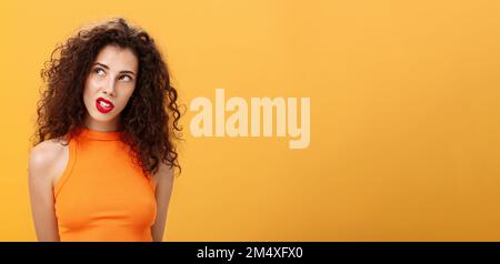 Élégante femme aux cheveux chauds et foncés avec boucles et rouge à lèvres dans un haut court tendance regardant dans le coin supérieur gauche intrigué et curieux de clenching dents Banque D'Images