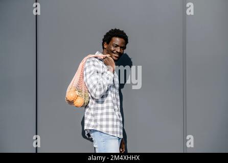 Homme souriant avec sac en maille debout devant le mur gris le jour de soleil Banque D'Images