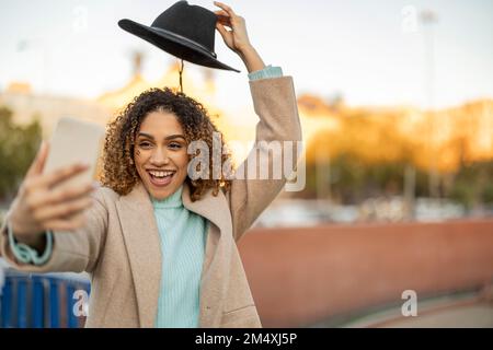 Une femme heureuse tenant un chapeau au-dessus de la tête prenant selfie à travers le smartphone Banque D'Images
