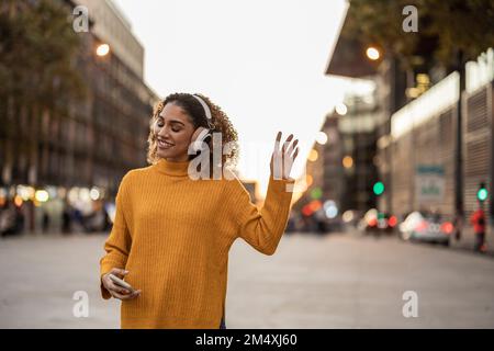 Femme souriante tenant un téléphone portable pour écouter de la musique à l'aide d'un casque sans fil et danser sur le sentier Banque D'Images