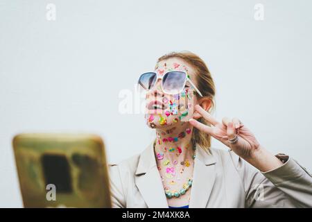 Femme avec des autocollants sur le visage gestant et prenant le selfie à travers le téléphone mobile en face de fond blanc Banque D'Images