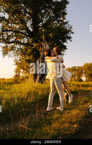 Photo verticale d'un jeune couple aimant embrassant les yeux fermés sur un pré vert en soirée d'été au coucher du soleil doré. Banque D'Images