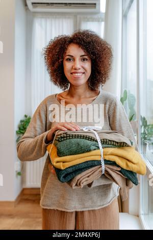 Jeune designer de mode heureux avec des vêtements pliés debout dans l'atelier Banque D'Images