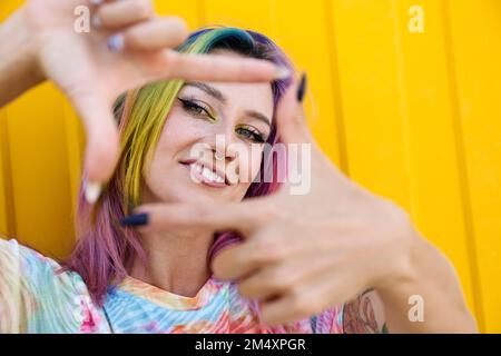 Jeune femme souriante faisant le cadre de doigt devant le mur jaune Banque D'Images