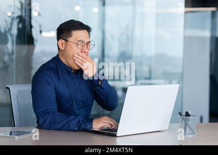 Un jeune asiatique fatigué en lunettes est assis dans le bureau à la table, travaillant sur un ordinateur portable. Il hurle, recouvre sa bouche avec sa main, s'endorme. Besoin de repos. Banque D'Images