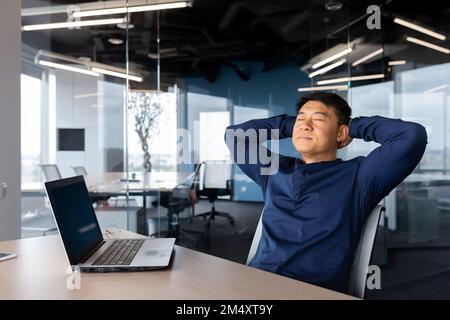 Pause café. Jeune homme d'affaires asiatique, indépendant assis au bureau sur une chaise. Il a mis ses mains derrière sa tête, fermé les yeux. Il repose, se détend, médite. Banque D'Images