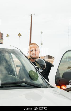 Image verticale d'un policier blanc de race blanche debout dans l'ouverture de la porte de sa voiture de patrouille avec des lumières sur la caméra. Banque D'Images