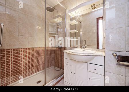 Petite salle de bains avec lavabo en porcelaine sur un meuble en bois blanc avec miroir intégré et cabine de douche avec écrans et carreaux de mosaïque Banque D'Images