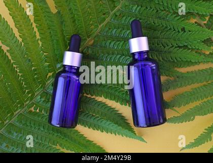 Flacons en verre bleu avec bouchon de pipette, vue de dessus. bouteille d'huile essentielle en verre avec pipette sur fond jaune avec feuilles tropicales. Maquette avec cosmétique Banque D'Images