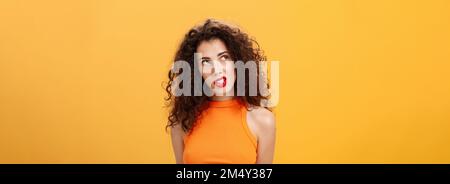 Élégante femme aux cheveux chauds et foncés avec boucles et rouge à lèvres dans un haut court tendance regardant dans le coin supérieur gauche intrigué et curieux de clenching dents Banque D'Images