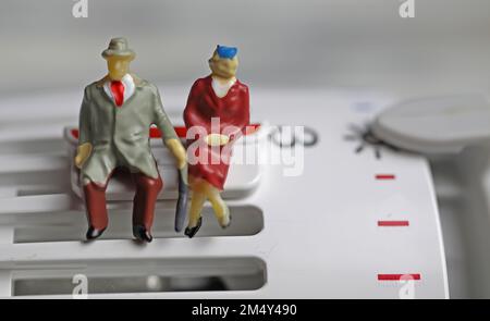 Figures de vieux couple retraité assis chaleureusement habillé sur un thermostat de chauffage à des températures froides sur clip de sauvegarde Banque D'Images