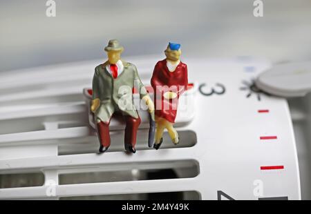 Figures de vieux couple retraité assis chaleureusement habillé sur un thermostat de chauffage à des températures froides sur clip de sauvegarde Banque D'Images