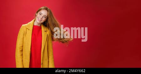 Charmante femme à tête rouge avec des taches de rousseur qui agite les cheveux en souriant large tête inclinable comme une coupe de cheveux volante dans l'air en posant ravie et insouciante sur le rouge Banque D'Images