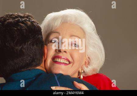 Une femme caucasienne aux cheveux gris plus âgée embrassant un jeune homme, elle sourit et est photographiée de derrière Banque D'Images