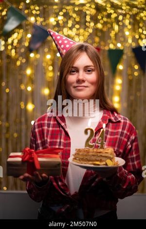 Fête d'anniversaire. Une jeune femme tient une assiette avec un savoureux gâteau d'anniversaire et un cadeau sur fond de fête. 24th anniversaire. Copier l'espace Banque D'Images