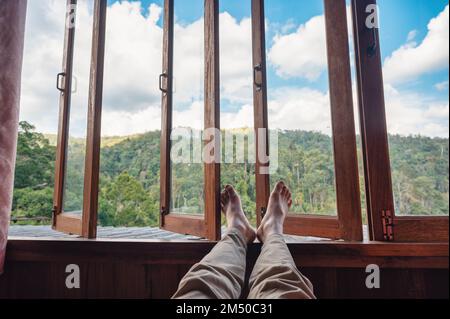 Pieds nus homme jambes de détente par fenêtre en bois dans la chambre et vue sur la montagne dans la campagne par jour ensoleillé Banque D'Images