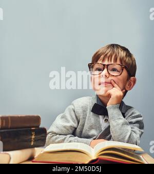 L'avenir appartient aux curieux. Studio photo d'un petit garçon intelligent lisant des livres et regardant réfléchi sur un fond gris. Banque D'Images