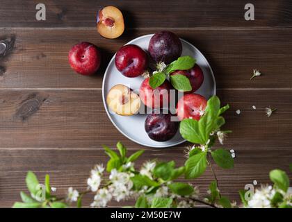 quelques prunes rouges juteuses dans une assiette bleue avec des fleurs de branches sur une serviette sur un fond de bois brun. Vue de dessus et image d'espace de copie Banque D'Images