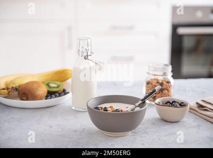 Le concept d'un petit déjeuner sain composé de yaourt végétarien, de granola et de fruits frais sur une table bleue sur fond blanc de cuisine. Vue avant. Banque D'Images