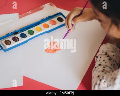 Un gros plan d'une fille qui peint un coeur sur papier blanc, en utilisant des aquarelles, le concept de la créativité et de l'éducation Banque D'Images