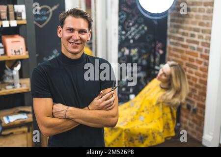 Professionnel heureux homme coiffeur dans le t-shirt noir tenant des ciseaux dans le salon de coiffure professionnel. Une cliente blonde portant une cape jaune assise à l'arrière-plan. Photo de haute qualité Banque D'Images