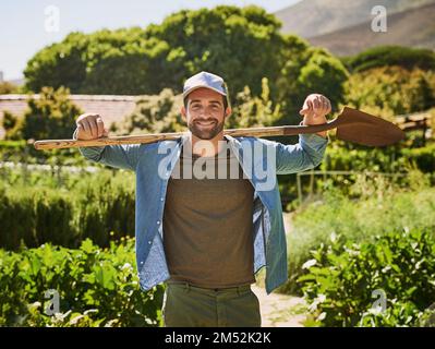 La vie est juste meilleure sur une ferme. Portrait d'un jeune agriculteur heureux tenant une bêche en posant dans les champs de sa ferme. Banque D'Images