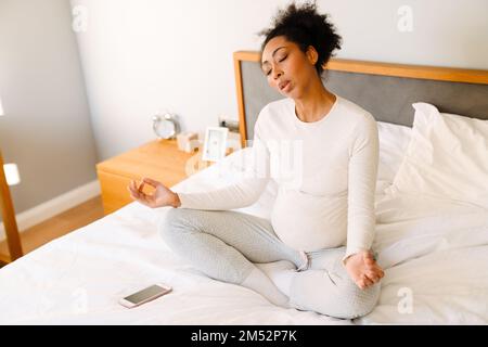 Jeune belle femme afro enceinte assise sur le lit avec les yeux fermés dans la pose de lotus et méditant dans la chambre confortable à la maison Banque D'Images