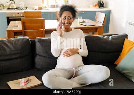 Jeune belle souriante heureuse femme afro enceinte assise sur un canapé et parlant sur son téléphone dans une pièce confortable à la maison Banque D'Images