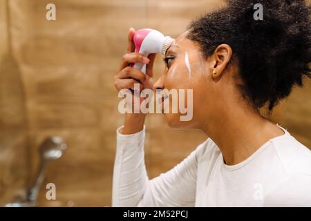 Concept beauté et soin de la peau. Jeune femme africaine utilisant de la mousse nettoyante pour laver son visage avec une brosse en silicone dans la salle de bains fermé Banque D'Images