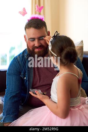 C'est un papa assez incroyable. une adorable petite fille qui met du maquillage sur son père à la maison. Banque D'Images